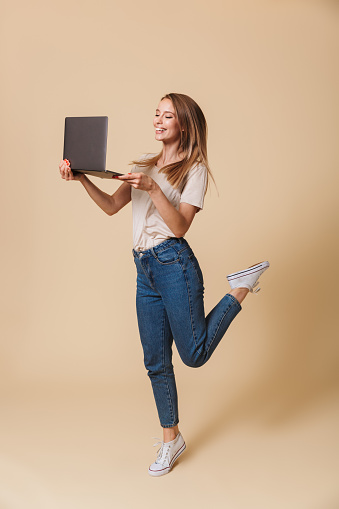Full length photo of smiling blond woman 20s wearing casual clothing lifting one leg while holding black laptop isolated over beige background