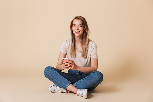 Portrait of a happy casual girl holding mobile phone while sitting on a floor with legs crossed isolated over beige background