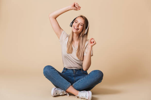 Pleased woman wearing casual clothing singing while listening to music via wireless earphones and sitting on floor with legs crossed, isolated over beige background Pleased woman wearing casual clothing singing while listening to music via wireless earphones and sitting on floor with legs crossed isolated over beige background isolated color photos stock pictures, royalty-free photos & images