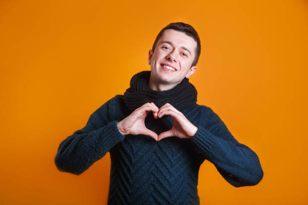 Smiling european student wearing a warm sweater and holding hands in heart gesture on yellow background stock photo