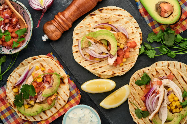 istock Mexican tacos with grilled chicken, avocado, corn kernels, tomato, onion, cilantro and salsa at black stone table. Traditional Mexican and Latin american street food. Top view. 1323884874