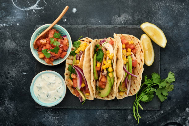 istock Mexican tacos with grilled chicken, avocado, corn kernels, tomato, onion, cilantro and salsa at black stone table. Traditional Mexican and Latin american street food. Top view. 1323885708