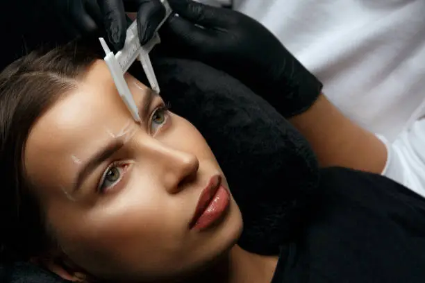 Woman in gloves makes measuring before permanent brow makeup to a glamorous brunette woman in beauty salon. Top view