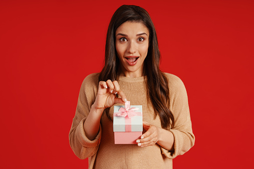 Attractive young woman in casual clothing holding a present and smiling while standing against red background