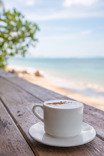 White cup of coffee on wooden tabletop on sand beach front of sea, close up. Copy space.