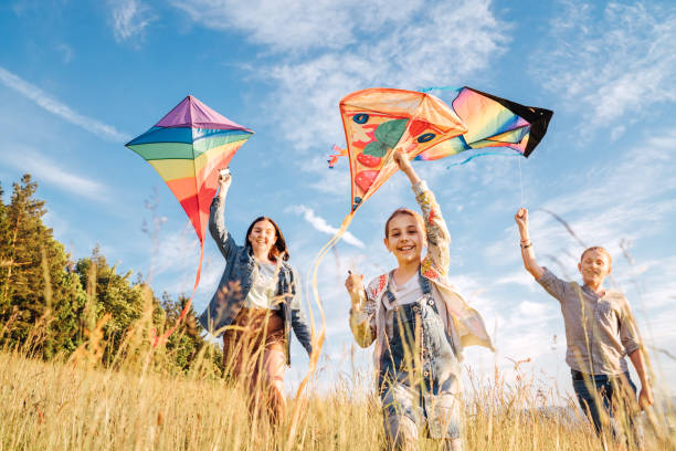 Smiling gils and brother boy running with flying colorful kites on the high grass meadow in the mountain fields. Happy childhood moments or outdoor time spending concept image. stock photo