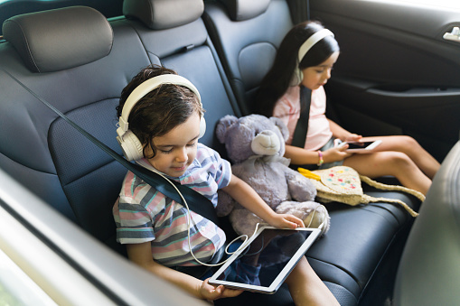 Beautiful kids watching a movie on a tablet with headphones while sitting in the car. Little siblings enjoying going with their parents on a trip