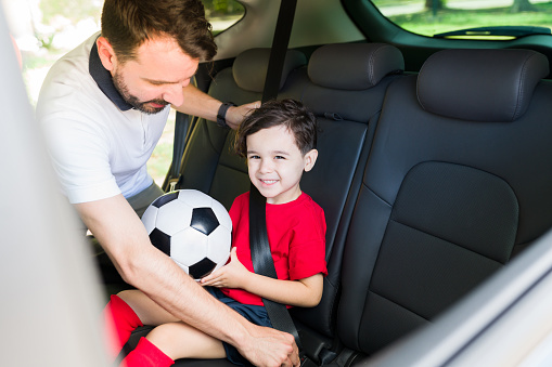 Cute little boy smiling and wearing a red uniform while holding a ball in the car. Caring dad putting the seat belt on to his son on their way to soccer practice