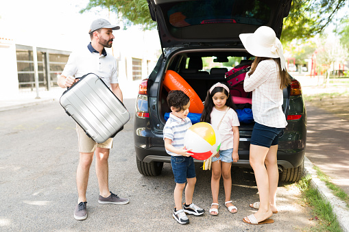 Hispanic parents packing their suitcases in the trunk of their car with their children. Young mom, dad and little kids ready to start a family vacation