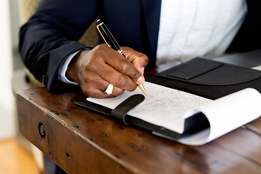 Close-up on mature African-American man’s hand working from home. He is wearing a navy blue jacket. He is sitting at the dinner table, white wall in the background. Horizontal close-up indoors shot with copy space. No face