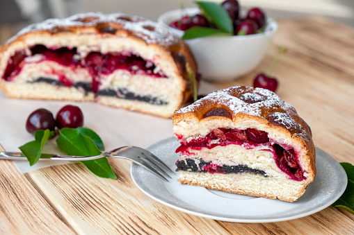 Piece of cherry pie on plate on rustic wooden board with full cake and sweet cherry berries on background. Easy recipe of berry dessert for everyday cooking. Homemade bakery by classic recipe.