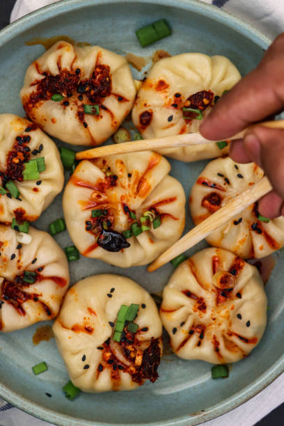 istock Full frame image of unrecognisable person using chopsticks to select steamed Momos (South Asian dumplings) from plate, white flour and water dough filled with pork and mixed vegetables, drizzled with chilli oil, elevated view 1323934033