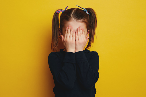 Cute little embarrassed girl having shy look, teen with ponytails covering her face with both hands, isolated over yellow background