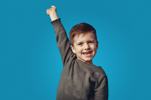 Excited schoolboy in casual wear standing with raised hand up on blue background while screaming and celebrating achievement