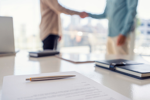 Two businessmen shaking hands with a contract. There is a pen on the contract document. Focus is on the foreground with the people in the office out of focus in the background. They are wearing casual business clothes.