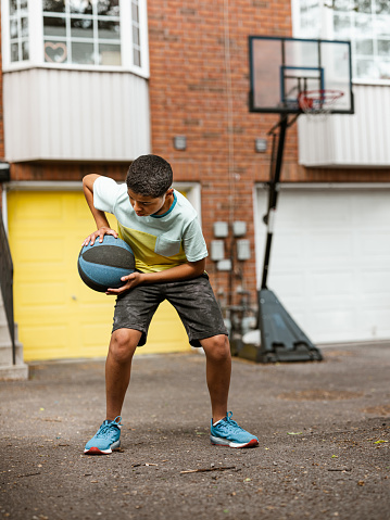 Mixed race boy playing basketball in the alley. He is wearing summer outfit with shorts and t-shirt. Exterior of urban townhouse lane in the city.