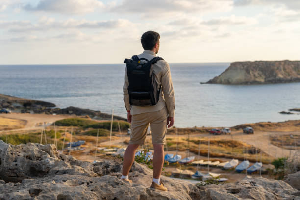 일몰에 키프로스에서 해안 아지오스 조지오스 페게이아스 근처 예로니소스 거룩한 섬의 아름다운 전망을보고 배낭과 남성 관광객. 지중해의 황량한 섬에서 거리를 바라보는 남자 등산객 - scotland cyprus 뉴스 사진 이미지