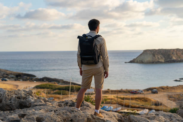 배낭을 가진 남자는 키프로스의 지중해 근처 일몰에 바위 해안을 따라 하이킹. 예로니소스(예로니소스) 또는 거룩한 섬의 전망을 감상하는 남성 관광객은 아지오스 조지오스 페게이아스 해안에 - scotland cyprus 뉴스 사진 이미지