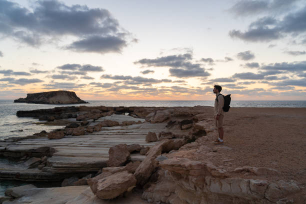 sea sunset view. man with backpack on rocks with beautiful view of yeronisos island near coast of agios georgios pegeias. guy enjoying ocean horizon, panoramic sunset in cyprus on mediterranean sea - scotland cyprus 個照片及圖片檔