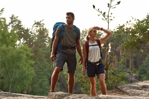 Cheerful young caucasian man and woman with large backpacks standing on rock while hiking in summer nature. Hiking, travel outdoor, recreation and active adventure concept