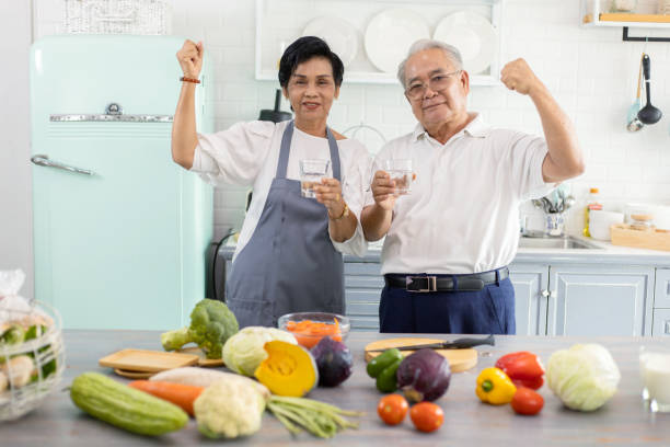Elderly Asian couple drinking water in the kitchen. The seniors have a smiling face and showing muscles of the arm while they are holding a glass of water. They are healthy and strong. stock photo
