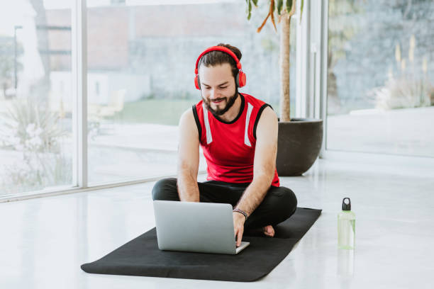 jeune homme latin avec des écouteurs rouges à la recherche de tutoriels vidéo de yoga sur internet assis sur un tapis - lotus position audio photos et images de collection