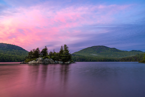 View of Lac Stukely at Parc National du Mont-Orford, Quebec, Canada during sunset. The sky is pink and purple. There is a little island on the lake. A chain of mountains is in the background.