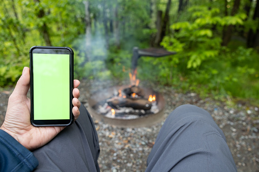 Close-up of Man Hands Reading on Chroma Key Green screen Mobile Phone by the Campfire. He could also be watching a video, video calling, reading emails, working, looking at social media, online shopping, etc… He is camping at Parc National du Mont-Orford.