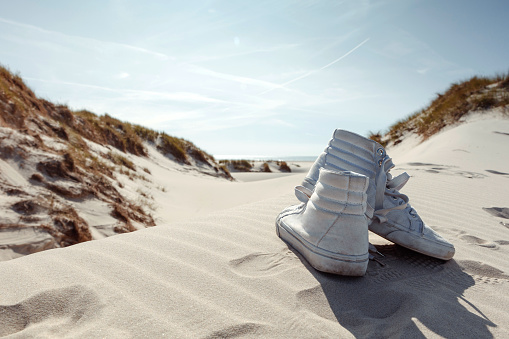Pair of white sneakers left on sand at beach. Dunes with green grass vegetation. Summer vacation, holiday at sea concept.