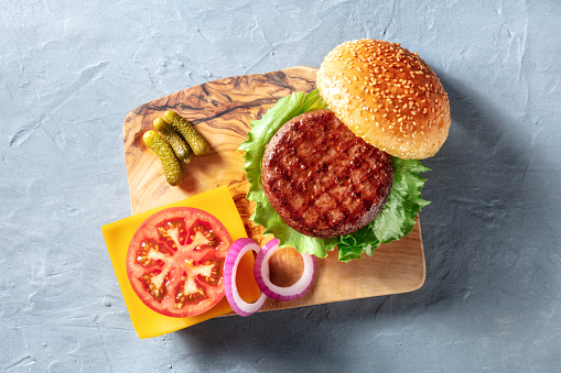 Burger ingredients, shot from the top on a wooden board. Beef patty with green salad, tomato, onion, Cheddar cheese and sesame buns, overhead shot