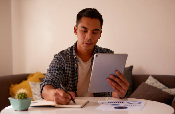 Shot of a handsome young man sitting alone and using a digital tablet to work from home I need to reschedule with this client book summary apps stock pictures, royalty-free photos & images