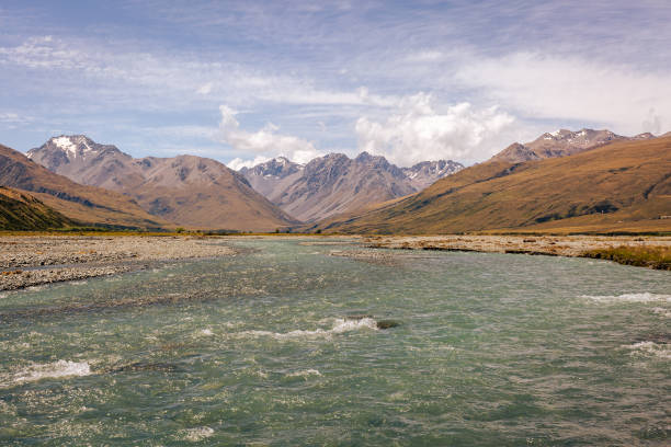 View of the shallow, swiftly flowing mountain river. In the background a mountain range with snow-covered peaks, white clouds in th esky