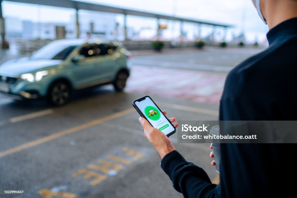 Chic young Asian man using mobile app device on smartphone to order a taxi pick up service by the urban road in downtown city street. Speedy and trustworthy service. Carsharing and business on the go themes Crowdsourced Taxi Stock Photo