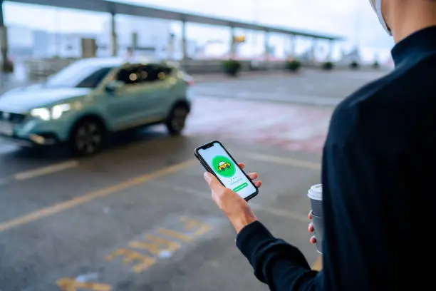 Photo of Chic young Asian man using mobile app device on smartphone to order a taxi pick up service by the urban road in downtown city street. Speedy and trustworthy service. Carsharing and business on the go themes