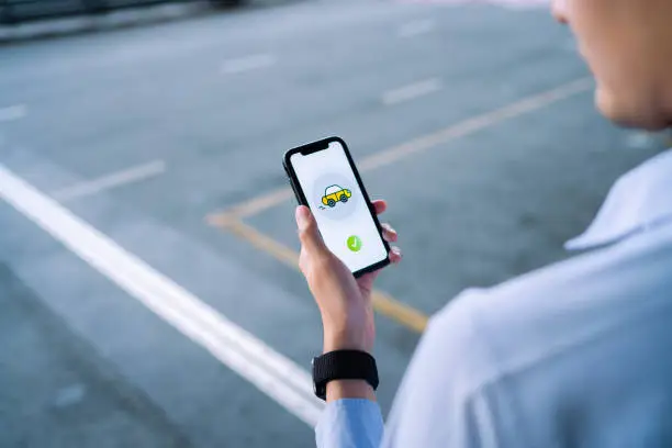 young Asian man ordering a taxi with mobile app on smartphone, waiting for taxi in downtown city street. Technology and transportation in everyday life