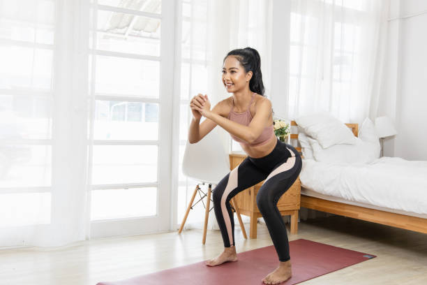 A beautiful Asian woman's fitness at home instead of going to the gym. She is doing squats on a yoga mat in the bedroom. She wears sportswear. Exercise concept for good shape stock photo