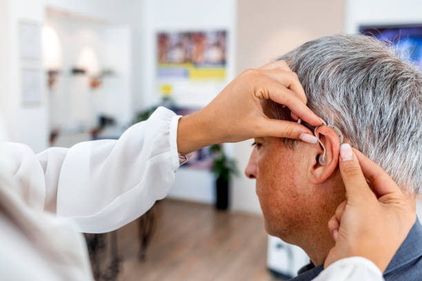 Female doctor fitting a male patient with a hearing aid Mature man at medical examination or checkup in otolaryngologist's office. Doctor Fitting To Male Patient Hearing Aid. hearing test stock pictures, royalty-free photos & images