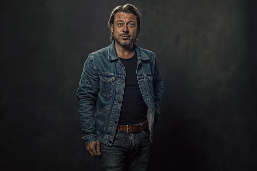 Blond man in jeans with a brown leather belt and a denim jacket in front of a dark grey wall.