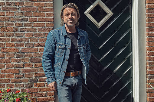 Blond man in jeans with a brown leather belt and a denim jacket in front of front door of an old farmhouse on a sunny day.