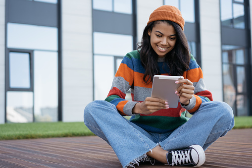 Happy student studying, learning language, online education concept. Beautiful African American woman using digital tablet, watching movie outdoors