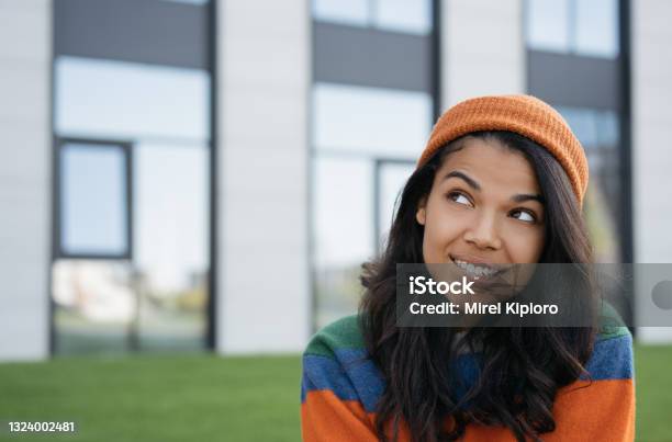 African American woman biting her lip, thinking, looking to copy space, standing outdoors. Close up portrait of young pensive hipster wearing casual clothes and orange hat