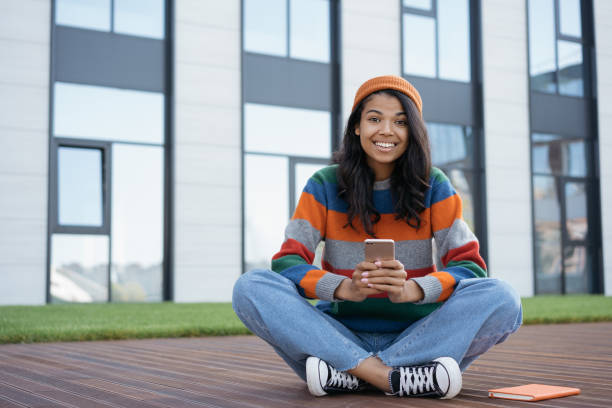 istock Happy emotional student studying, holding smartphone, online education concept. Beautiful African American woman using mobile phone, chatting, looking at camera, sitting in park 1324002545