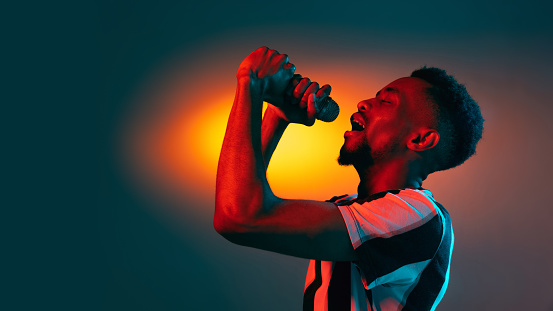 Singing, performing. African-american young man's portrait on red studio background in neon light. Beautiful male model rapping. Concept of human emotions, facial expression, youth, sales, ad.