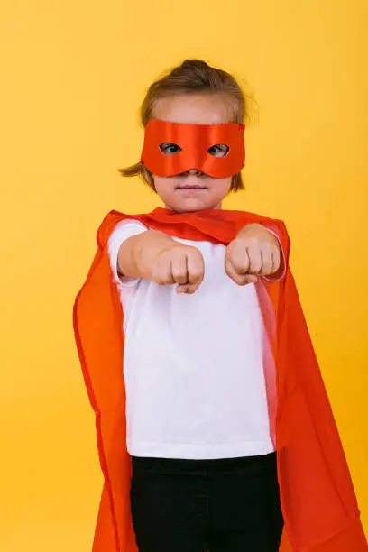 Little blonde girl dressed as a superheroine superhero with a red cape and mask, with arms in a flying position, on a yellow background