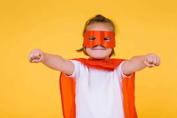 Little blonde girl dressed as a superheroine superhero with a cape and red mask, smiling with open arms in a flying position, on yellow background