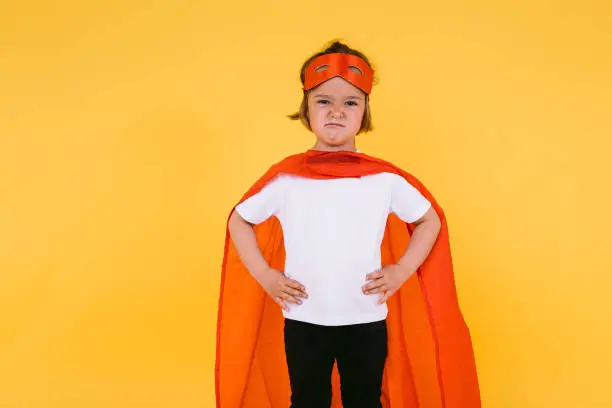 Little blonde girl dressed as a superheroine superhero with a cape and red mask, angry, with her arms akimbo serious, on yellow background