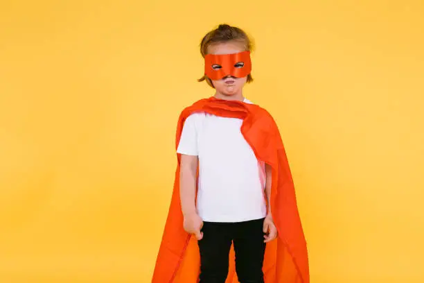Little blonde girl dressed as a superheroine superhero with a cape and red mask, angry, with the cape to the side, on yellow background