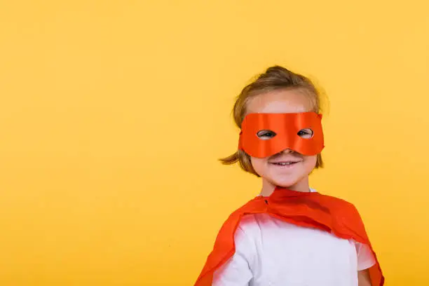 Little blonde girl dressed as superheroine superhero with cape and red mask, smiling, on yellow background