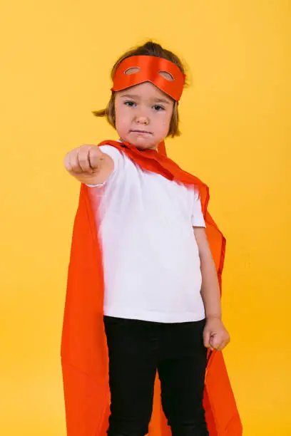 Little blonde girl dressed as a superheroine superhero with a cape and red mask, raising her arm in a flying position, on yellow background