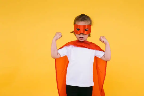 Little blonde girl dressed as a superheroine superhero with a red cape and mask, squeezing her arms as a sign of strength, on yellow background